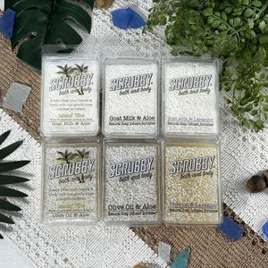 May include: Six white plastic containers with clear soap bars inside. Each container has a label that says "Scrubby bath and body" and lists a different scent: Island Vibe, Goat Milk & Aloe, Goat Milk & Lavender, Olive Oil & Aloe, Olive Oil & Lavender.