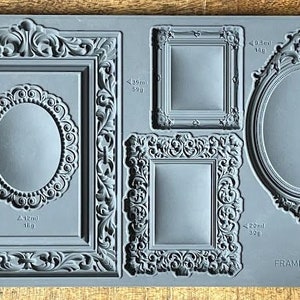 May include: A silicone mold for making decorative frames. The mold features four different frame designs, including a round frame, a square frame, and two ornate frames. The mold is gray and has measurements in milliliters and grams.
