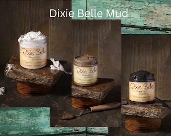 May include: Three jars of Dixie Belle Mud in white, brown, and black, each labeled 8 oz | 237 ml. The jars are displayed on stone platforms, with a small metal spatula resting near the brown jar, showcasing the thick, textured paste consistency.