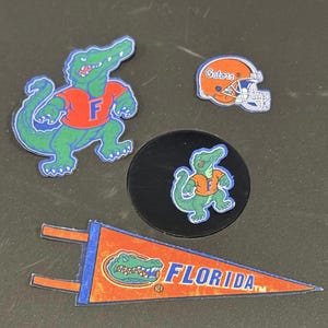 May include: A collection of Florida Gators themed stickers and a pennant. The stickers include a green alligator wearing an orange shirt with a blue outline, a football helmet, and a circular sticker with the alligator. The pennant reads "FLORIDA".