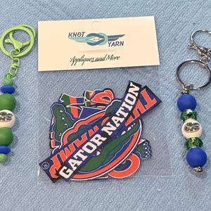 May include: Two beaded keychains with metal rings and a clear plastic bag containing several fabric patches. The left keychain has green and blue beads with a logo bead. The right keychain has blue and green beads with a logo bead. The patches feature the text GATOR NATION and alligator graphics.