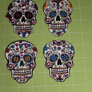 May include: Four colorful sugar skull designs with intricate floral patterns and bright colors, including blue, orange, yellow, red, and purple.