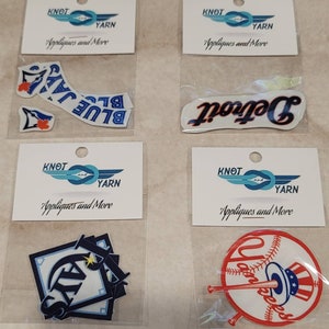 May include: Four packages of iron-on patches featuring logos for the Detroit Tigers, New York Yankees, Toronto Blue Jays, and Seattle Mariners. Each package includes a Knot and Yarn logo with the text "Appliques and More".