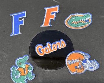 Op de afbeelding: Diverse patches en een cirkelvormig embleem met het Florida Gators-logo. Bevat de letter "F" in blauw en oranje, een Gator-mascotte en een voetbalhelm. Het woord "Gators" is ook afgebeeld.