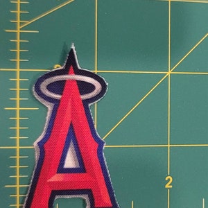 May include: A red and blue fabric patch with the letter "A" in a stylized font, surrounded by a halo. The patch is on a green cutting mat.