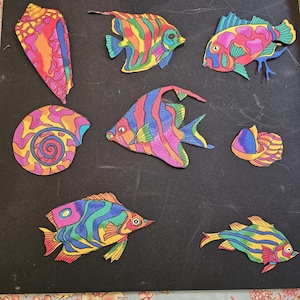May include: A collection of colorful paper cutouts of seashells and fish. The fish are striped with rainbow colors and have black outlines. The seashells are pink, purple, and yellow with swirls.