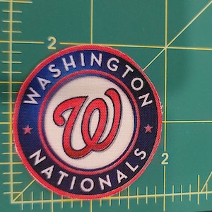 May include: A red, white, and blue fabric patch with the Washington Nationals logo. The logo features a white "W" with a red outline inside a blue circle with white stars and the words "Washington Nationals" around the circle.