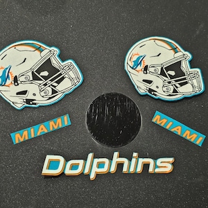May include: Two white and blue Miami Dolphins football helmet stickers with the team name and "Miami" written below each helmet. A black circle sticker with the team name "Dolphins" written below it.