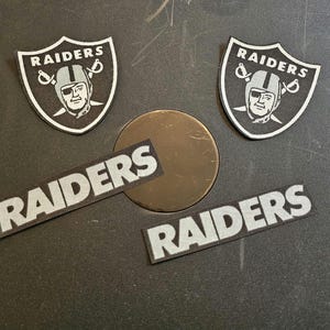 May include: An assortment of Raiders-themed patches and a circular metal object. Two shield-shaped patches feature the Raiders logo with a helmeted face and crossed swords. Two rectangular patches spell out "RAIDERS" in white letters. The metal object is bronze.