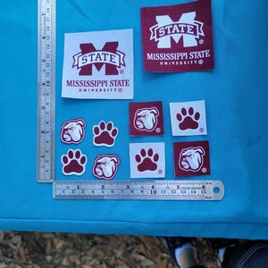 May include: Nine iron-on patches featuring the Mississippi State University logo, a bulldog mascot, and a paw print. The patches are maroon and white and are arranged on a blue surface.
