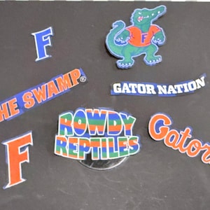 May include: A collection of colorful University of Florida Gators-themed stickers and decals. Includes the word "Gators" in orange script, a green alligator mascot, and phrases like "The Swamp" and "Rowdy Reptiles."