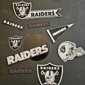 May include: A collection of nine black and white iron-on patches featuring the Raiders logo and the word "Raiders".