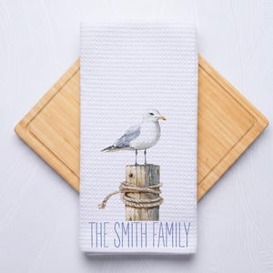 May include: White waffle-weave kitchen towel featuring a watercolor illustration of a seagull perched on a wooden piling. The towel has the text "THE SMITH FAMILY" in blue script at the bottom. The towel is on a wooden cutting board.