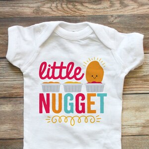 以下が含まれることがあります： ピンク、オレンジ、ターコイズ、黄色の「little NUGGET」の文字が入った白いベビーボディスーツ。笑顔のチキンナゲットの漫画がデザインされています。ボディスーツは半袖で、下部にスナップが付いています。