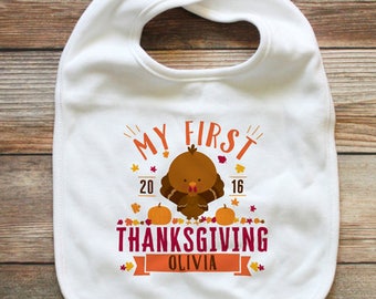 Puede incluir: Un babero blanco para bebé con un gráfico de un pavo marrón, dos calabazas naranjas y hojas de otoño coloridas. El texto dice MY FIRST THANKSGIVING 2016 y OLIVIA en una cinta. El diseño incluye letras en color naranja, rojo y rosa oscuro.