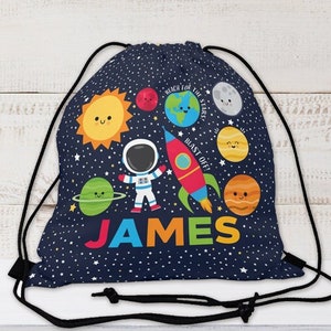 May include: A navy blue drawstring bag with a space theme. It features a cartoon astronaut, planets, stars, and a rocket with the text "Reach for the stars! Blast off!" and the name "JAMES" in colorful letters.