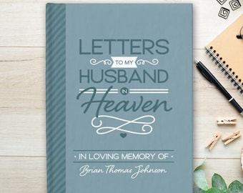 May include: A blue journal titled "Letters to My Husband in Heaven" with the name "Brian Thomas Johnson" below. The cover has a striped spine and decorative text. The journal is on a wooden surface with a pen, notepad, and other stationery.