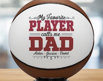May include: A brown basketball with a white panel featuring the text "My Favorite PLAYER calls me DAD" in maroon. The names Aiden, Jessica, and David are below the text. The ball is on a black stand.