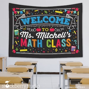 May include: A black classroom banner with the words "WELCOME TO Ms. Mitchell's MATH CLASS" in colorful letters. The banner has a chalkboard design with school-themed illustrations like apples, calculators, and pencils. The banner is hanging above several school desks.