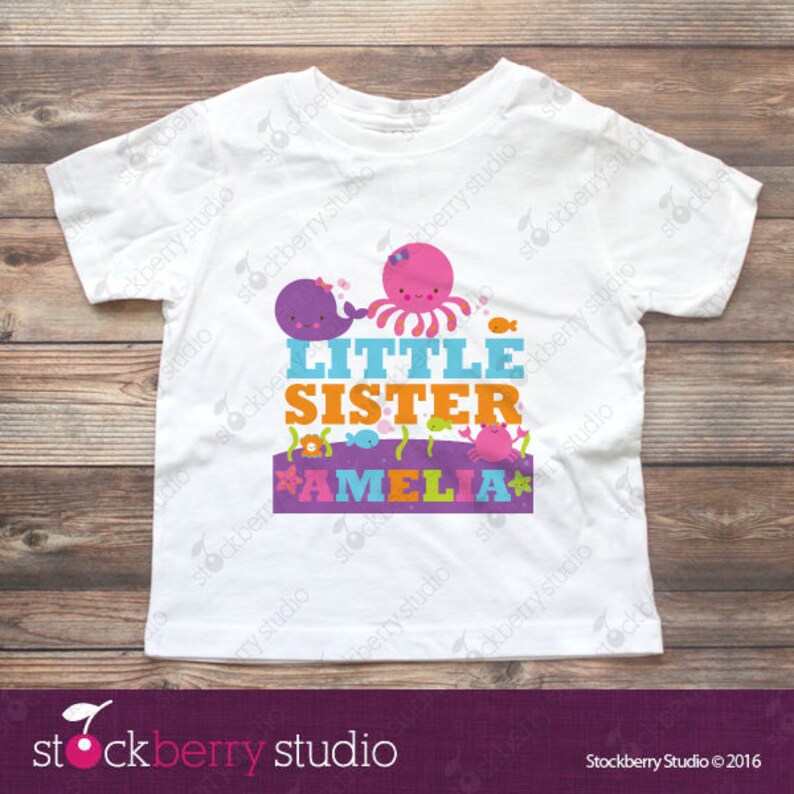 Puede incluir: Una camiseta blanca de manga corta con un gr&aacute;fico colorido de una ballena morada, un pulpo rosa y un cangrejo rosa. El texto dice LITTLE SISTER en azul y naranja, con el nombre AMELIA impreso en rosa sobre un fondo de pancarta de color morado.