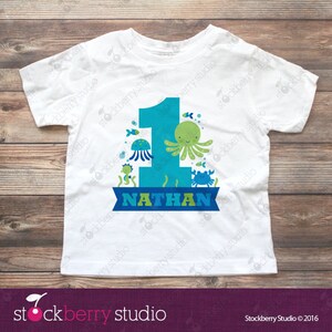 Puede incluir: Una camiseta blanca de manga corta con un gr&aacute;fico grande del n&uacute;mero uno en azul. El dise&ntilde;o incluye ilustraciones de un pulpo verde, una medusa azul, un cangrejo azul y un caballito de mar. Una banda azul en la parte inferior muestra el nombre NATHAN en verde.