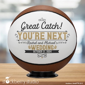 May include: A white basketball with brown leather panels, decorated with black and gold text that reads "Great Catch! You're Next Rachel and Michael's Wedding October 21, 2024".