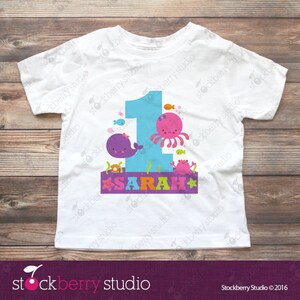 Puede incluir: Una camiseta blanca de manga corta con un gr&aacute;fico grande del n&uacute;mero uno en azul claro. El dise&ntilde;o incluye una ballena morada, un pulpo rosa, un cangrejo y peces peque&ntilde;os. El nombre SARAH est&aacute; impreso en letras de colores sobre una banda morada.