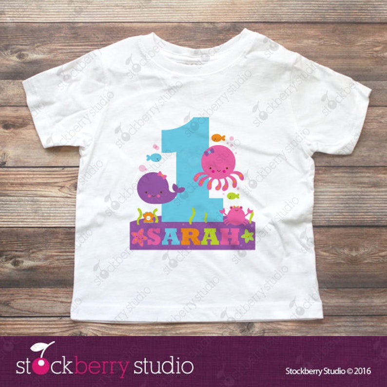 Puede incluir: Una camiseta blanca de manga corta con un gr&aacute;fico grande del n&uacute;mero uno en azul claro. El dise&ntilde;o incluye una ballena morada, un pulpo rosa, un cangrejo y peces peque&ntilde;os. El nombre SARAH est&aacute; impreso en letras de colores sobre una banda morada.