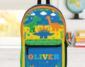 May include: A colourful backpack featuring a dinosaur theme. The backpack has a blue front with the name "OLIVER" in orange, green, and blue letters. The top of the backpack has a design with green, orange, and blue dinosaurs.