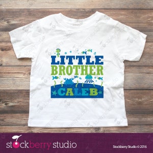 Puede incluir: Una camiseta blanca de manga corta con un gr&aacute;fico azul y verde que incluye el texto LITTLE BROTHER CALEB. El dise&ntilde;o presenta ilustraciones de un caballito de mar, peces, un cangrejo, una medusa y estrellas de mar alrededor del texto.