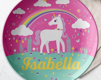 May include: A pink and green children's plate with a white unicorn standing under a rainbow. The plate has a green border and the name "Isabella" written in yellow letters.