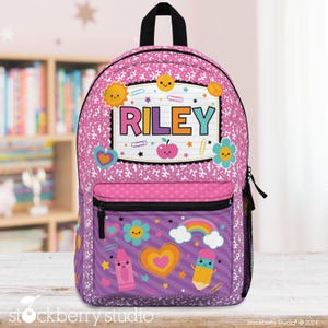May include: Pink and purple backpack with the name "RILEY" in a marquee-style design. The backpack features a front pocket with a rainbow, crayon, and pencil graphic. The main body has a repeating pattern and school-themed illustrations.
