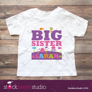 Puede incluir: Una camiseta blanca de manga corta con el texto BIG en morado y SISTER en rosa. Debajo, una banda morada muestra el nombre SARAH en letras multicolores, rodeado de ilustraciones coloridas de un cangrejo, una medusa y peces peque&ntilde;os.