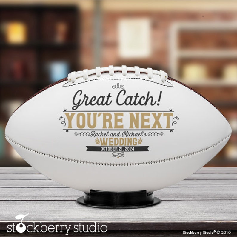 Pode incluir: Bola de futebol branca com costuras pretas e um gr&aacute;fico preto e dourado que diz "Great Catch! You're Next" com o texto "Rachel and Michael's Wedding October 21, 2024" abaixo.