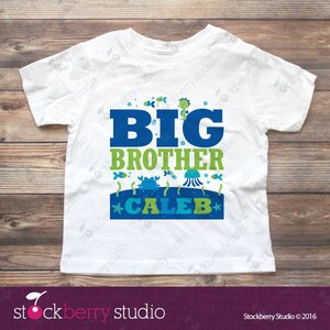 Puede incluir: Una camiseta blanca de manga corta con un estampado gr&aacute;fico que incluye el texto BIG BROTHER CALEB en azul y verde. El dise&ntilde;o presenta ilustraciones de un cangrejo, una medusa, un caballito de mar y peces peque&ntilde;os sobre un fondo azul marino.