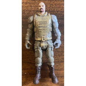 Peut inclure: Une figurine d'action militaire avec une tête chauve et une moustache. La figurine porte un gilet tactique vert et gris, un pantalon et des bottes. Les bras et les mains de la figurine sont gris. La figurine est debout.