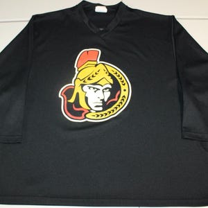 May include: Black hockey jersey with a v-neck and long sleeves. The jersey features a large team logo on the front, depicting a stylized face in yellow, red, and white.