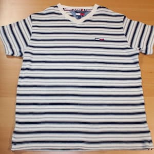 Pode incluir: Uma camisa de manga curta com decote em V listrada. A camisa apresenta listras horizontais em branco, azul marinho e cinza. A gola tem a inscrição "TOMMY JEANS". Um pequeno logotipo está no peito esquerdo.