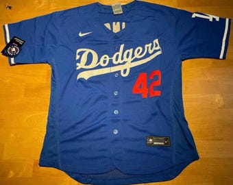 May include: A blue Dodgers baseball jersey with white lettering and the number 42 in red. The jersey has a button-up front, short sleeves, and a Nike logo. Tags are attached to the sleeve.