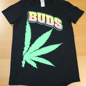 May include: Black t-shirt with a large, light green cannabis leaf graphic on the left side. The word "BUDS" is printed across the top in a gradient of pink and yellow with a green outline. The shirt is made of a soft material.