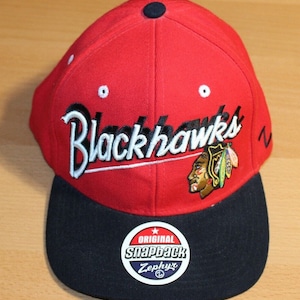 Könnte beinhalten: Rote und schwarze Baseballkappe mit dem Schriftzug "Blackhawks" in weiß auf der Vorderseite. Die Kappe hat einen schwarzen Schirm und ein schwarzes Logo mit einem indianischen Kopf und Federn auf der rechten Seite. Auf der Rückseite der Kappe befindet sich ein weißes "Zephyr"-Logo mit einem Stern und den Worten "Original Snapback".