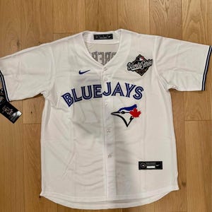 以下が含まれることがあります： 白地の野球ユニフォーム。青と赤のアクセントが施されています。「BLUEJAYS」の文字が胸に青い筆記体で書かれ、鳥とカエデの葉のロゴ、そして「World Series」のパッチが付いています。ナイキのロゴが見えます。