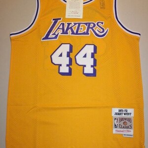 May include: A yellow basketball jersey with purple and white trim. The jersey has "LAKERS" written across the chest in purple and the number "44" in white. The jersey has a tag that says "1971-72 JERRY WEST HARDWOOD CLASSICS Mitchell & Ness".