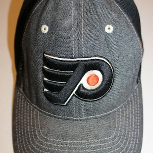 Puede incluir: Gorra de béisbol de mezclilla gris con el logotipo de los Flyers de Filadelfia. El logotipo es negro con detalles en blanco y naranja. La gorra tiene una parte trasera de malla y una visera curva con costuras blancas.