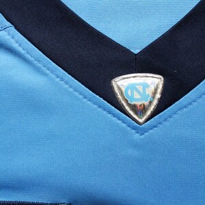 May include: A light blue jersey with a navy blue collar and a silver patch with a blue and white logo on the front.