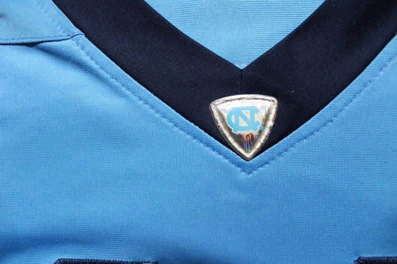 May include: A light blue jersey with a navy blue collar and a silver patch with a blue and white logo on the front.