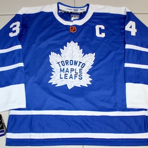 May include: A blue Toronto Maple Leafs hockey jersey with white accents. The jersey features the number 34 on the sleeves, a white maple leaf logo with "TORONTO MAPLE LEAFS" text, and a "C" for captain. The jersey has a white collar and white stripes.