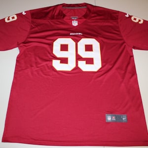 Puede incluir: Una camiseta de fútbol burdeos con el número 99 en blanco en la parte delantera y en las mangas. La camiseta tiene ribetes dorados en las mangas y una etiqueta adjunta. La palabra "REDSKINS" está encima del número.
