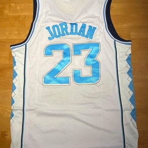 May include: White basketball jersey with blue and navy trim. The back of the jersey has the name "JORDAN" and the number "23" in blue with a white outline.