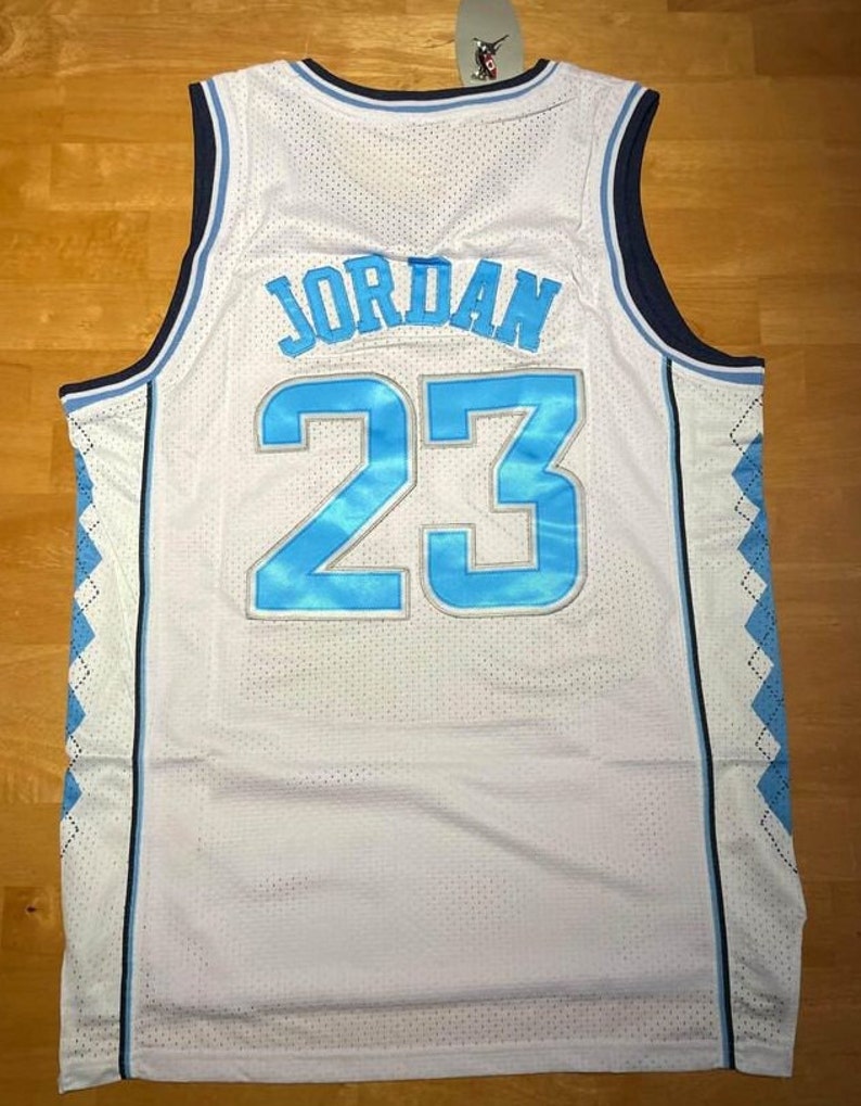 May include: White basketball jersey with blue and navy trim. The back of the jersey has the name "JORDAN" and the number "23" in blue with a white outline.